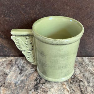 May include: A light green ceramic mug with a decorative handle. The mug has a distressed, vintage look.
