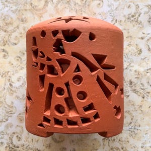 May include: A cylindrical terracotta candle holder with geometric cutouts. The holder has a rounded top with star-shaped openings and a series of circular and angular cutouts along the sides. The color is a warm reddish-brown.