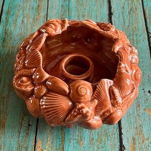 May include: A terracotta candle holder shaped like a seashell with a raised design of various seashells. The holder has a central opening for a candle.