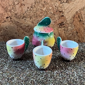 May include: A set of four ceramic teacups and a teapot, all shaped like a cactus with green, pink, yellow, and white colors. The cups have black dots and the teapot has a green leaf on top.