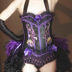 May include: Black and purple corset with decorative gold accents, purple bows, and beaded fringe. The corset features a halter neck and is paired with black gloves and a feather skirt. A burlesque-style costume.