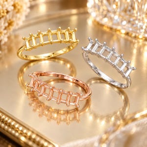 May include: Three metal ring settings in gold, silver, and rose gold tones. Each ring features a thin band and a top section with five rectangular, open-frame prongs designed to hold stones. The rings are arranged in a cluster on a reflective surface.