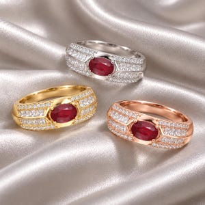 May include: Three rings featuring oval-cut red centre stones surrounded by rows of small clear accent stones. The rings are crafted in silver-tone, gold-tone, and rose gold-tone metal bands, each showcasing a wide, multi-row design with pavé settings.