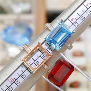 May include: A silver ring with a rectangular light blue gemstone, a rose gold ring setting without a stone, and a loose rectangular red gemstone, all positioned on a metal ring mandrel marked with numerical size measurements.