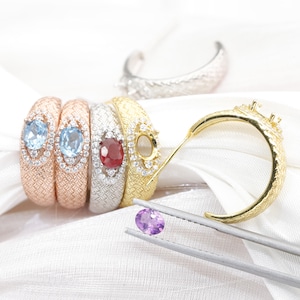 Könnte beinhalten: Eine Auswahl an Edelsteinringen in Roségold, Silber und Gold, jeweils mit einem strukturierten Band. Die Ringe zeigen ovale Edelsteine in Blau, Rot und eine Fassung für einen Stein. Eine Pinzette hält einen violetten Edelstein.