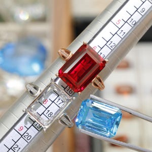 May include: A rose gold ring with a large rectangular red gemstone, a silver ring setting without a stone, and a loose rectangular blue gemstone held by tweezers, all positioned on a ring mandrel showing size 16 to 18 measurements.