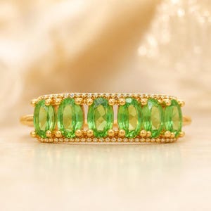 May include: A gold ring featuring six oval-cut, vibrant green gemstones arranged in a horizontal row. The band and the settings are crafted from polished gold, with small, clear, round accent stones lining the top and bottom edges of the setting.