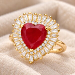 May include: A gold ring featuring a large heart-shaped red centre stone. The stone is surrounded by a halo of small round clear stones, followed by an outer border of baguette-cut clear stones arranged in a sunburst pattern on a gold band.