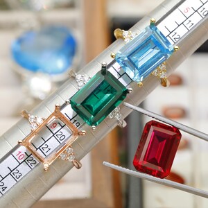 May include: Three rings with emerald-cut stones in light blue, green, and a rose-gold setting, positioned on a ring mandrel with a numbered scale. A loose, rectangular red gemstone is held by metal tweezers next to the rings on the mandrel.