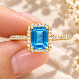 Könnte beinhalten: Ein Goldring mit einem rechteckigen, smaragdgeschliffenen, leuchtend blauen Edelstein in der Mitte, umgeben von einem Kranz aus kleinen, runden, weißen Steinen. Die Ringschiene ist zudem mit einer Reihe kleiner, runder, weißer Steine besetzt.