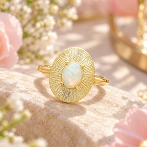 May include: A gold-toned ring featuring an oval-shaped iridescent opal cabochon set in the center. The setting is a textured, sunburst-inspired oval plate with radiating lines extending from the stone to the outer edge of the metal frame.
