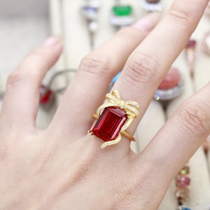 May include: A gold ring featuring a large, rectangular, deep red gemstone as the centerpiece. The setting is adorned with a delicate gold bow motif encrusted with small, clear, sparkling stones that frame the top and sides of the red stone.