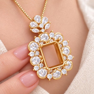 May include: A gold-toned pendant necklace featuring a central rectangular empty setting surrounded by a cluster of round and pear-shaped clear white stones. The pendant hangs from a delicate gold-toned box chain, showcasing a detailed metal frame.