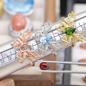 May include: Three flower-shaped rings in rose gold, silver, and gold, each with a gemstone center. A loose, teardrop-shaped red gemstone is held by tweezers. The rings are on a ring sizer.
