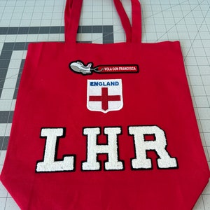 Puede incluir: Bolsa de lona roja con letras bordadas blancas "LHR" y un pequeño parche de avión con el texto "Vola con Francesca" y un parche de la bandera de Inglaterra.