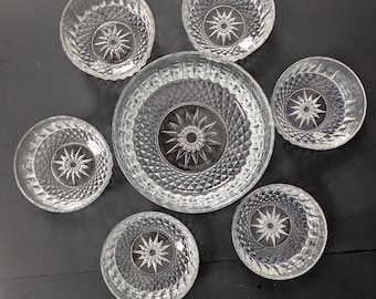 May include: A set of seven clear glass bowls. The bowls have a diamond pattern and a starburst design in the center. One large bowl is surrounded by six smaller bowls, all on a dark surface.