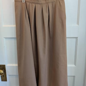 May include: A solid tan midi-length skirt featuring a high waistband with a single button closure on the side and vertical pleats extending from the waist down to the hem, creating a structured and flared silhouette in a smooth fabric.