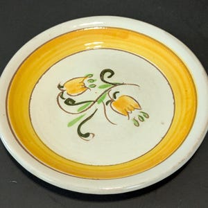 May include: A small, round ceramic plate with a white center and a yellow border. The center features a floral design with yellow flowers and green leaves. The plate is likely used for serving small portions of food.