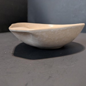 May include: A beige ceramic bowl with a unique, asymmetrical shape. The bowl has a smooth, matte finish and a slightly curved rim. The interior is a lighter shade of beige. The bowl is sitting on a dark surface.