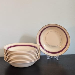 May include: A set of nine cream-colored ceramic bowls stacked together, with one additional bowl displayed upright on a clear plastic stand. Each bowl features a wide, dark maroon band encircling the interior rim and a thin gold-tone line.