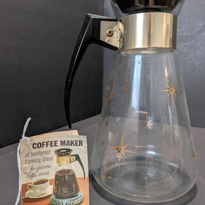 May include: Vintage glass coffee maker with a black handle and gold accents. The clear glass carafe features a starburst design. A tag reads "COFFEE MAKER of heatproof Corning Glass for gracious coffee service."