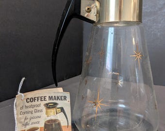 Puede incluir: Cafetera de vidrio vintage con asa negra y detalles dorados. La jarra de vidrio transparente presenta un diseño de estrella. Una etiqueta dice "COFFEE MAKER of heatproof Corning Glass for gracious coffee service."