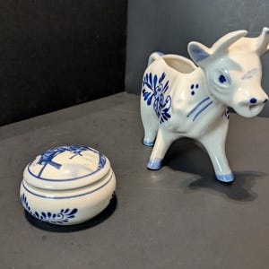 May include: A white ceramic cow creamer and a matching lidded trinket box, both adorned with blue floral and geometric patterns. The cow creamer has a detailed face and stands on four legs. The trinket box is oval-shaped.
