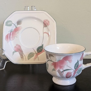 May include: A white ceramic teacup and matching square saucer featuring a delicate floral pattern with pink flowers and green leaves. Both pieces have a thin grey rim, and the teacup is designed with a pedestal base and a curved handle.