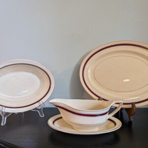 May include: A set of cream-colored ceramic tableware featuring a dark maroon band around the edges. The collection includes an oval serving platter, a smaller oval bowl, and a gravy boat with an attached saucer, all displaying a consistent design.