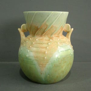 May include: A ceramic vase in green and tan hues. The vase features a rounded body, two handles, and a flared rim. The surface is adorned with small white dots and a textured design, suitable for home decor.