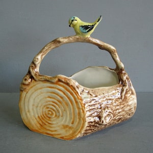 May include: A ceramic log-shaped basket with a handle, topped with a yellow and blue bird. The basket is painted in shades of brown and cream, with a textured finish to resemble wood. The end of the log has a circular design.