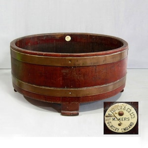May include: A round, dark brown wooden tub featuring two horizontal brass bands and three short feet. A small circular white label on the side reads A. LISTER & CO. LTD. MAKERS DURSLEY ENGLAND, with a small metal pin in the centre of the label.