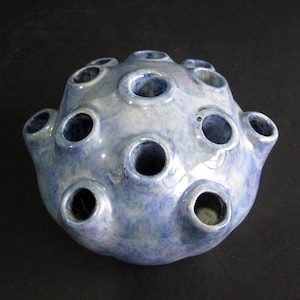 May include: A spherical ceramic flower frog with a glossy, mottled blue and white glaze. The object features multiple circular openings distributed across its rounded surface, designed to hold individual flower stems in an upright position.