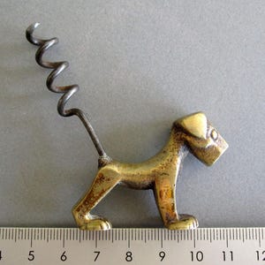 May include: A brass-coloured metal corkscrew shaped like a standing dog with a dark, spiralled metal worm extending from its back. The dog figure is positioned above a ruler, showing a length of approximately 9 cm from nose to tail.