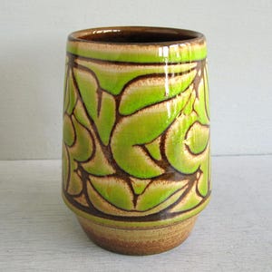 May include: A cylindrical ceramic vase with a glossy finish. The vase features a pattern of green leaf-like shapes outlined in brown against a beige background. The rim and base are brown.