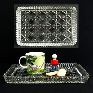 May include: Two rectangular, clear glass trays with diamond-patterned centres and textured edges. One tray holds a mug with a cartoon design, a small figurine, and two biscuits. The other tray is empty.