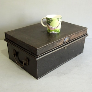 May include: A dark brown metal storage box with a hinged lid and a handle. A ceramic mug with a green and white design sits on top of the box. The mug features a cartoon character relaxing in a hammock.
