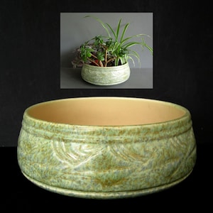 May include: A round ceramic planter with a light green mottled glaze and a textured leaf pattern around the exterior. The interior of the bowl is a smooth, solid beige colour. The planter has a low, wide profile with a thick, rounded rim.