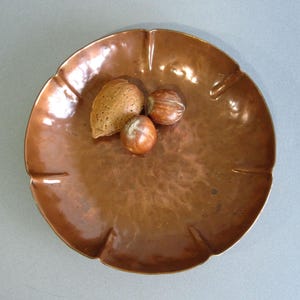 May include: A round, hammered copper bowl with a scalloped edge and a reflective metallic finish. Inside the bowl, there are three whole nuts, including one large almond in its shell and two smaller, round brown hazelnuts resting on the surface.