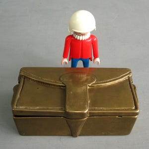 May include: A brass-coloured treasure chest with a clasp, and a Playmobil figure wearing a white helmet, red jacket, and blue trousers. The chest is rectangular with a curved top, set against a grey background.