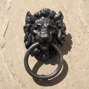 May include: A detailed, dark metal lion head door knocker with a circular handle. The lion's face is intricately sculpted, with a prominent mane and a ring-shaped handle. The knocker is mounted on a light-coloured stone surface.