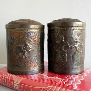 May include: Two antique metal canisters with lids. One canister features an embossed elephant design, the other depicts figures. The canisters are cylindrical and rest on a red book with gold lettering.