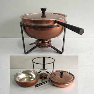 May include: A copper chafing dish with a lid, a long black handle, and a black metal stand featuring a burner component. The set includes a separate copper bowl with a silver-toned interior, a matching copper lid, and a black metal frame stand.