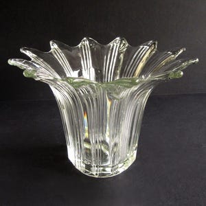 May include: Clear glass vase with a flared, scalloped rim. The vase has vertical ridges and a wide opening, resembling a flower. The glass is transparent, allowing light to pass through, and sits on a dark surface.