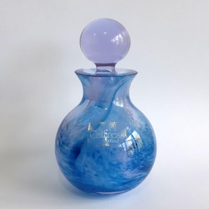 May include: A Caithness Scotland glass perfume bottle with a rounded body featuring a swirling blue and white marbled pattern. The bottle has a clear glass neck and a spherical, solid light purple glass stopper resting on top.