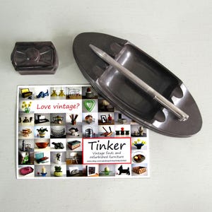May include: A dark grey rectangular lidded box and a matching dark grey oval-shaped tray with a central handle. A rectangular card is included featuring a grid of various product images, the text "Love vintage?" and "Tinker Vintage finds and refurbished furniture".