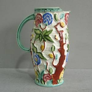 May include: A ceramic jug featuring a white textured surface with raised, hand-painted floral and tree motifs. The design includes a brown tree trunk, green leaves, and flowers in shades of red, blue, and yellow. It has a light green handle.