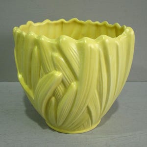 May include: A pale yellow ceramic planter featuring a textured exterior with overlapping, leaf-like vertical ridges. The vessel has a wide, scalloped rim and a rounded body that tapers towards a circular base, showcasing a uniform matte finish.