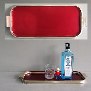 May include: A rectangular serving tray with a metallic silver frame and a textured red interior surface. The tray features integrated side handles. A clear glass tumbler, a small toy figure, and a blue glass bottle of Bombay Sapphire gin sit on it.