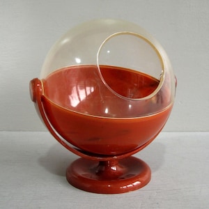 May include: A retro, red-orange plastic terrarium with a clear, domed lid. The terrarium has a circular opening on the side and stands on a round base. The design includes curved handles on either side of the base.